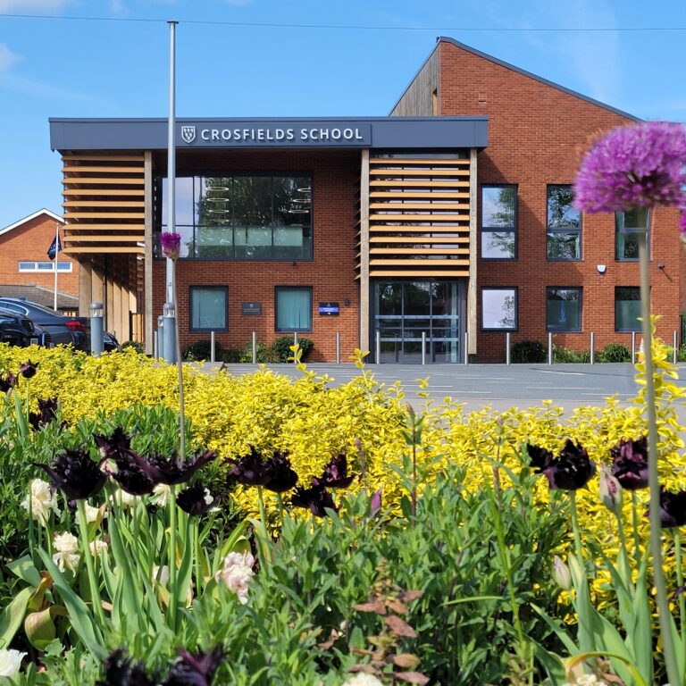 Exterior view of Crosfields School with brick building and yellow flower beds in the foreground under a blue sky.