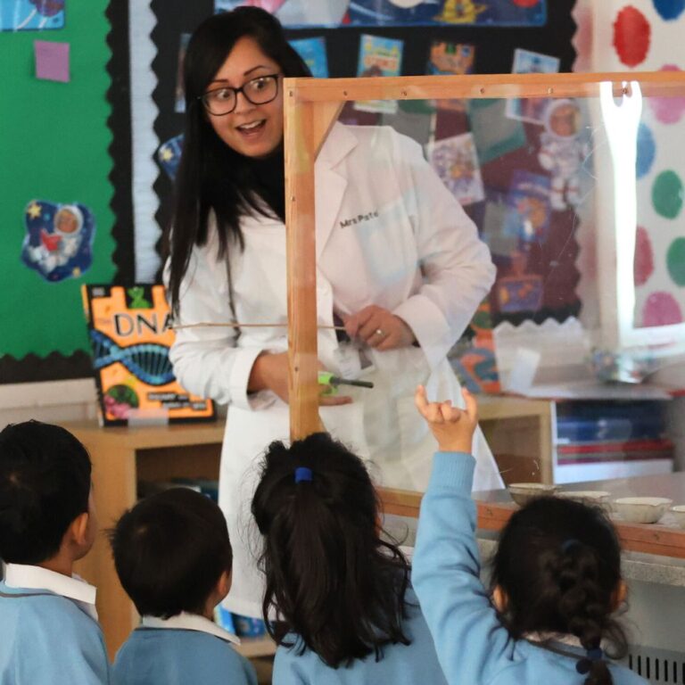 Science teacher in a white lab coat demonstrates a science activity to a group of children in blue uniforms, gathered around a clear protective enclosure in a colorful classroom.
