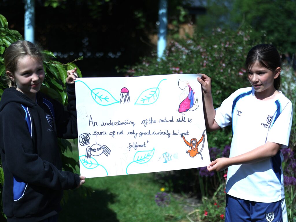 Two girls in school uniforms hold a large poster with drawings of leaves, animals, and text outdoors in a garden setting.