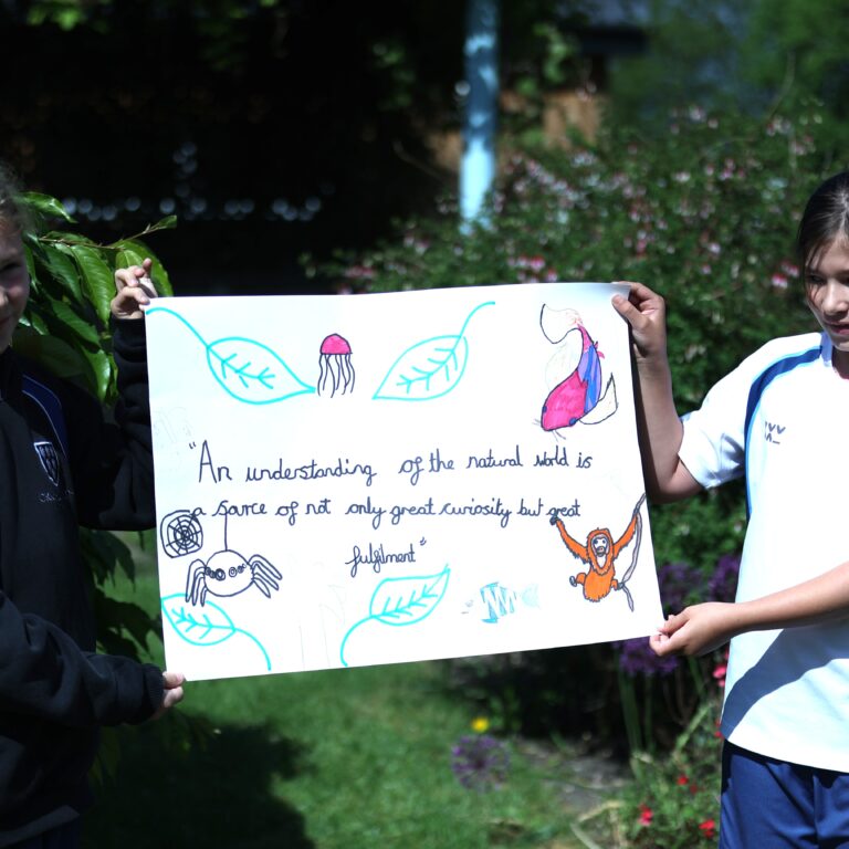 Two girls in school uniforms hold a large poster with drawings of leaves, animals, and text outdoors in a garden setting.