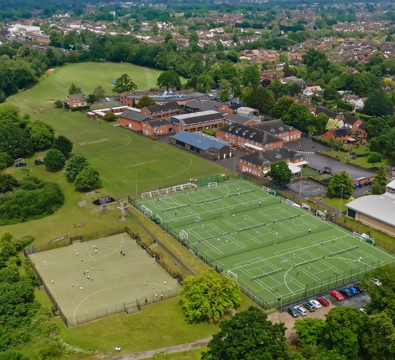 Aerial view of a school campus with soccer fields, tennis courts, and a gym building amid trees and houses nearby.
