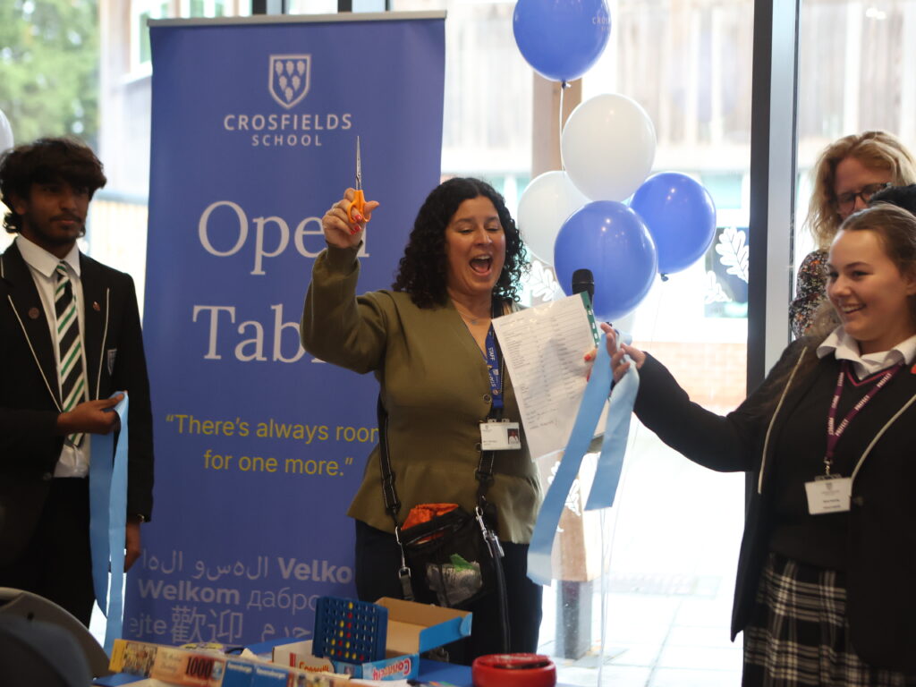 A group of students and staff celebrating a school event near a blue Crossfields banner with balloons. A woman holds scissors and a sheet of paper, smiling.