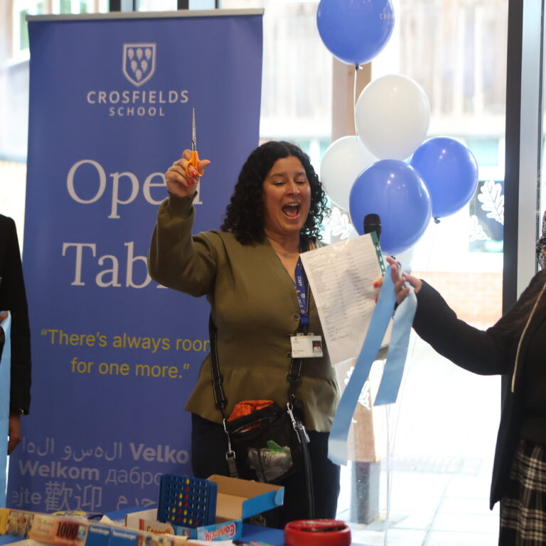 A group of students and staff celebrating a school event near a blue Crossfields banner with balloons. A woman holds scissors and a sheet of paper, smiling.