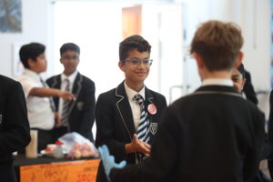 Smiling student in a blazer and striped tie with glasses, standing among peers at a school event.