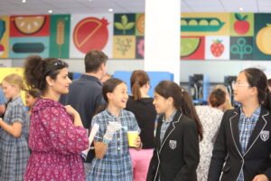 Group of students in plaid uniforms chat and laugh in a crowded indoor school setting, with a woman in a purple patterned dress nearby.