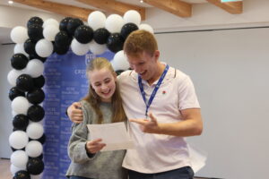 Two people smile as they hold a paper together at a GCSE results day event, with a staff member guiding them, and a black-and-white balloon arch behind.