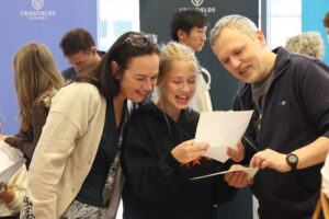 Three people (an adult woman, a young girl, and an adult man) lean in and smile as they read a document together at a school setting with a Crossfields banner in the background.