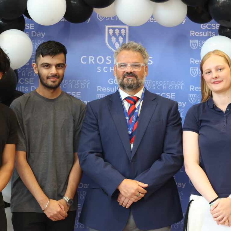 Four students posing with a staff member in front of a blue school backdrop and a black-and-white balloon arch at a school event.