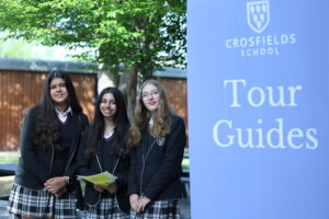 Three smiling female students in school uniforms stand beside a tall blue Crosfields School Tour Guides sign.
